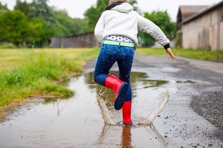 Happy kid boy wearing red rain boots and walking during sleet and rain on rainy cloudy day. Child in colorful casual clothes jumping into puddle. Having fun outdoors, healthy children activityの写真素材