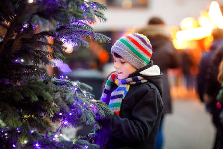 Little cute kid boy having fun on traditional Christmas market during strong snowfall. Happy child enjoying traditional family market in Germany. Schoolboy standing by illuminated xmas tree.の写真素材