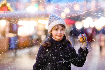 Cute little kid girl having fun on traditional Christmas market during strong snowfall. Happy child eating traditional curry sausage called wurst. schoolgirl standing by illuminated xmas tree.の写真素材