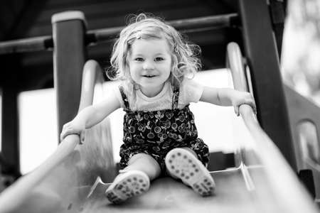 Cute toddler girl playing on slide on outdoor playground. Beautiful baby in colorful shorts trousers having fun on sunny warm summer day. Child sliding downの写真素材