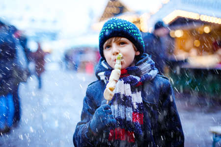Little cute kid boy eating white chocolate covered fruits on skewer on traditional German Christmas market. Happy child on traditional family market in Germany during snowy day.の写真素材