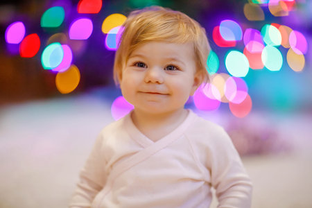 Adorable baby girl holding colorful lights garland in cute hands. Little child in festive clothes decorating Christmas tree with family. First celebration of traditional holiday called Weihnachtenの写真素材