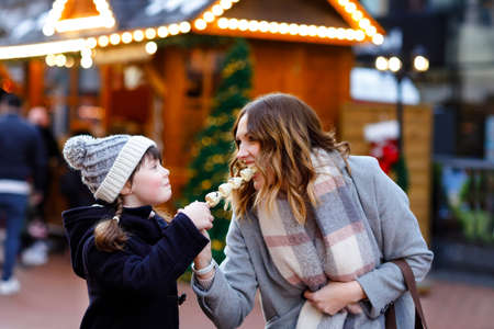 Mother and daughter eating white chocolate covered fruits and strawberry on skewer on traditional German Christmas market. Happy girl and woman on traditional family market in Germany during snowy dayの写真素材