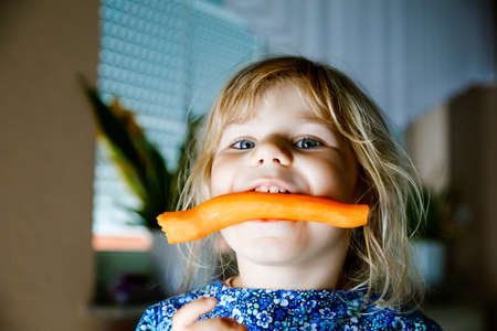 Cute adorable toddler girl holding and biting into fresh carrot. Beatuiful child having healthy snack. Smiling happy kid eating bio organic vegetablesの写真素材