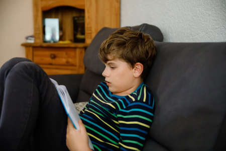 Cute blond little kid boy reading magazine or book in domestic room. Excited child reading loud, sitting on couch. Schoolkid, family, education. Homeschooling during covid timeの写真素材