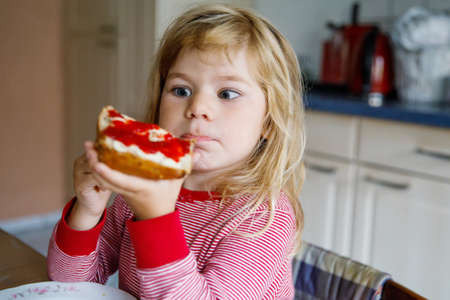 Cute funny toddler girl eats sweet bun for breakfast. Happy child eating bread roll with strawberry jam. Health food for children and kids with selfmade jellyの写真素材