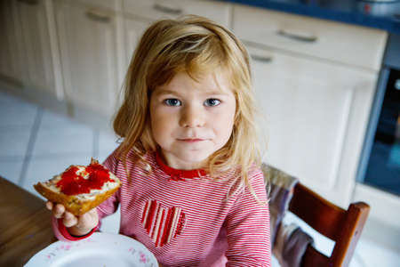 Cute funny toddler girl eats sweet bun for breakfast. Happy child eating bread roll with strawberry jam. Health food for children and kids with selfmade jellyの写真素材