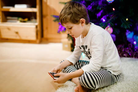 Cute little blond kid boy using smartphone on Christmas with decorated tree on background. Happy healthy hild having fun at home. Popular gift for xmasの写真素材