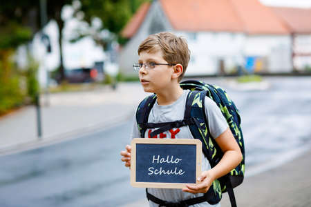 Happy little kid boy with glasses and backpack or satchel. Schoolkid on the way to middle or high school. Child outdoors on the street. Back to school. On desk Hello school in German.の写真素材