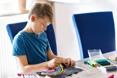 Little school kid boy paint rainbow with different stick colors on black background during pandemic...