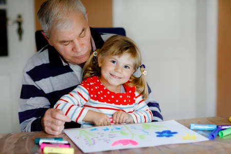Cute little baby toddler girl and handsome senior grandfather painting with colorful felt pens and pencils at home. Grandchild and man having fun together, creative family.の写真素材