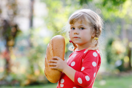 Little toddler girl holding big loaf of bread. Funny happy child biting and eating healthy bread,...