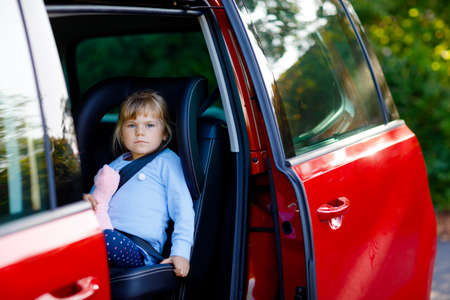 Adorable toddler girl sitting in car seat, holding plush soft toy and looking out of the window on nature and traffic. Little kid traveling by car. Child safety on the road. Family trip and vacationsの写真素材