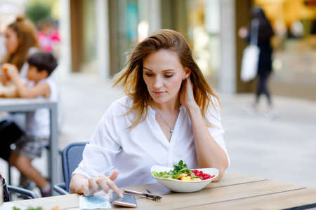 Young beautiful woman eating fresh salad in outdoor restaurant. Woman enjoying lunch bowl with fresh vegetables. Healthy food. On summer day.の写真素材
