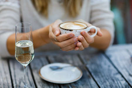 Closeup of hands of woman drinking cappuccino and cup of coffee and glass champagne while waiting for friend and breakfast.の写真素材