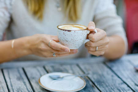 Closeup of hands of woman drinking cappuccino and cup of coffee while waiting for friend and breakfast. Happy alone woman in outdoor cafe or restaurant on sunny summer day.の写真素材