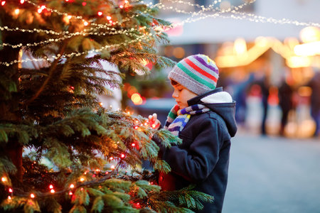 Little cute kid boy having fun on traditional Christmas market during strong snowfall. Happy child enjoying traditional family market in Germany. Schoolboy standing by illuminated xmas tree.の写真素材