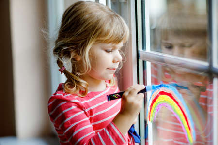 Adorable little toddler girl with rainbow painted with colorful window color during pandemic quarantine. Child painting rainbows and hearts around the world with words Lets all be well.の写真素材