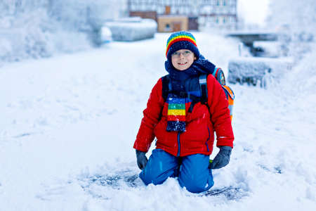 Gorgeous little school kid boy with glasses of elementary class walking to school during strong snowfall. Snowy streets in city. Child with backpack or satchel in colorful winter clothesの写真素材