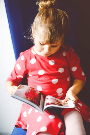 Little stylish toddler girl reading and looking fashion magazine or catalogue. Adorable child having...