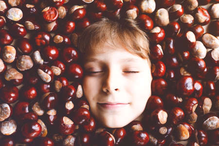 Funny portrait of preteen or school kid boy with lots of chestnuts. Smiling happy child having fun on autumn day with a school project.の写真素材