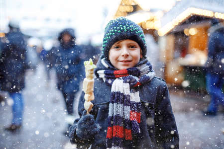 Little cute kid boy eating white chocolate covered fruits on skewer on traditional German Christmas market. Happy child on traditional family market in Germany during snowy day.の写真素材