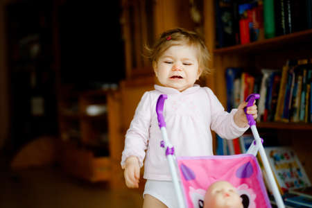 Cute adorable baby girl making first steps with doll carriage. Beautiful toddler child pushing stroller with toy at home. Happy daughter learning walking and standingの写真素材
