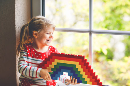 Cute little toddler girl by window create rainbow with colorful plastic blocks during pandemic...