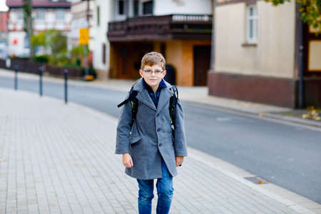 Happy kid boy with glasses and backpack or satchel. Schoolkid in stylish fashon coan on the way to middle or high school on cold autumn day. Healthy child outdoors on the street, on rainy day.の写真素材