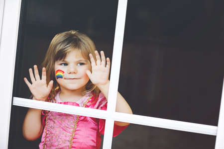 Cute lonely little toddler girl in princess dress sitting by window with rainbow with colorful colors on face during pandemic coronavirus quarantine. Children make and paint rainbows around the worldの写真素材