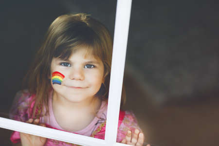 Cute lonely little toddler girl in princess dress sitting by window with rainbow with colorful colors on face during pandemic coronavirus quarantine. Children make and paint rainbows around the worldの写真素材