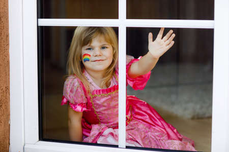 Cute lonely little toddler girl in princess dress sitting by window with rainbow with colorful colors on face during pandemic coronavirus quarantine. Children make and paint rainbows around the worldの写真素材