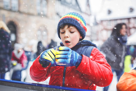 Happy little kid boy in colorful warm clothes on skating rink of Christmas market or fair drinking...