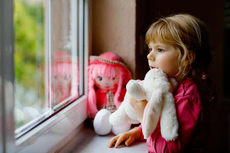 Cute toddler girl sitting by window and looking out on rainy day. Dreaming child with doll and soft toy feeling happy. Self isolation concept during corona virus pandemic time. Lonely kid.の写真素材