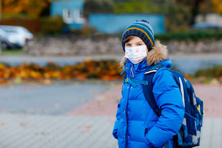 Little kid boy wearing medical mask on the way to school. Child backpack satchel. Schoolkid on cold autumn or winter day with warm clothes. Lockdown and quarantine time during corona pandemic diseaseの写真素材