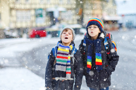Two little kids boys of elementary class walking to school during snowfall. Happy children having fun and playing with first snow. Siblings ans friends with backpack in colorful winter clothes.の写真素材