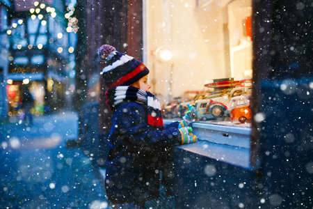 Cute toddler boy looking on car toys in a window on Christmas time season. Fascinated child in winter clothes dreaming and wishing. Window decorated with xmas gifts. Snow falling down, snowfall.の写真素材