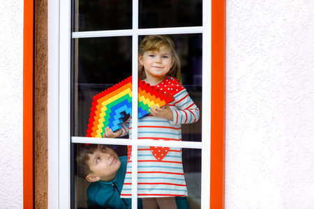 Toddler girl and kid boy by window create rainbow with colorful plastic blocks during pandemic...