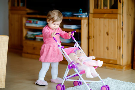 Cute adorable baby girl make first steps and push with doll carriage. Beautiful toddler child...