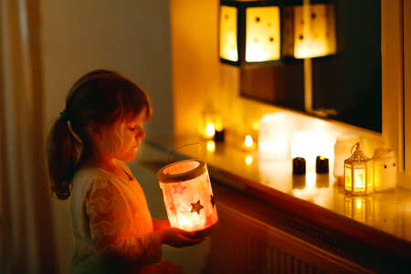Little kid girl sitting by window with selfmade hand crafted lanterns with candles for St. Martin procession. Toddler child looking at glowing lantern. German tradition Martinsumzug. Home decorationの写真素材