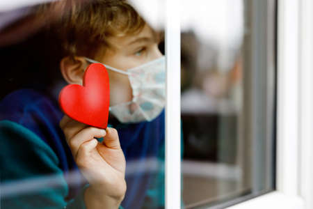 Lovely little school kid boy by a window wearing medical mask and holding wooden heart during pandemic coronavirus quarantine. Lonely upset child, self isolation concept.の写真素材