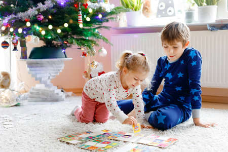 Two little chilren, cute toddler girl and school kid boy playing together card game by decorated Christmas tree. Happy healthy siblings, brother and sister having fun together. Family celebrating xmasの写真素材