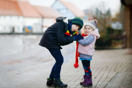 School kid boy and brother warming hands of little sister, toddler girl on cold snowy grey winter day. Family, two lovely siblings children playing with snow, outdoors during snowfall.の写真素材