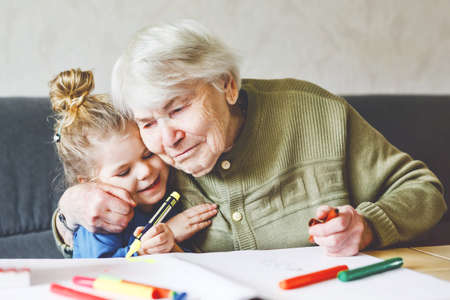 Beautiful toddler girl and grand grandmother drawing together pictures with felt pens at home. Cute child and senior woman having fun together. Happy family indoorsの写真素材
