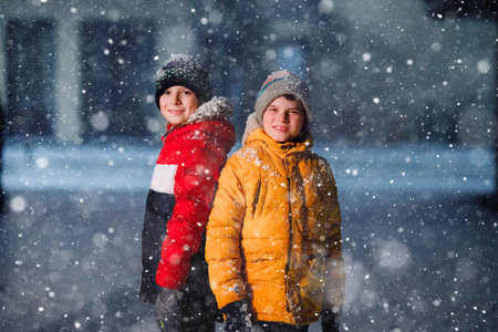 Two little kids boys of elementary class walking to school during snowfall on early dark morning....