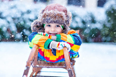 Little kid boy enjoying sleigh ride during snowfall. Happy preschool kid riding on vintage sledge....
