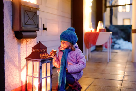 Little cute toddler girl with with a big light candle lantern. Happy child on Christmas decorating backyard of house. Family holiday, illumination, lights and candles.の写真素材