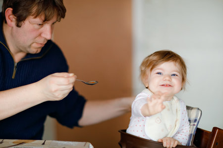 Father feeding baby girl from spoon mashed vegetables and puree. food, child, feeding and people concept -cute toddler, daughter and dad with spoon sitting in highchair and eating at home.の写真素材