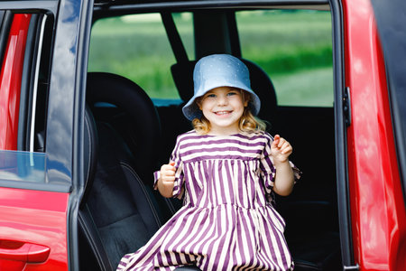Adorable toddler girl sitting in car seat and looking out of the window on nature and traffic. Little kid traveling by car. Child safety on the road. Family trip and vacations in summerの写真素材