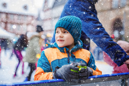 Happy little kid boy in colorful warm clothes on skating rink of Christmas market or fair drinking hot punch or chocolate. Healthy child having fun on ice skate. people having active winter leisureの写真素材